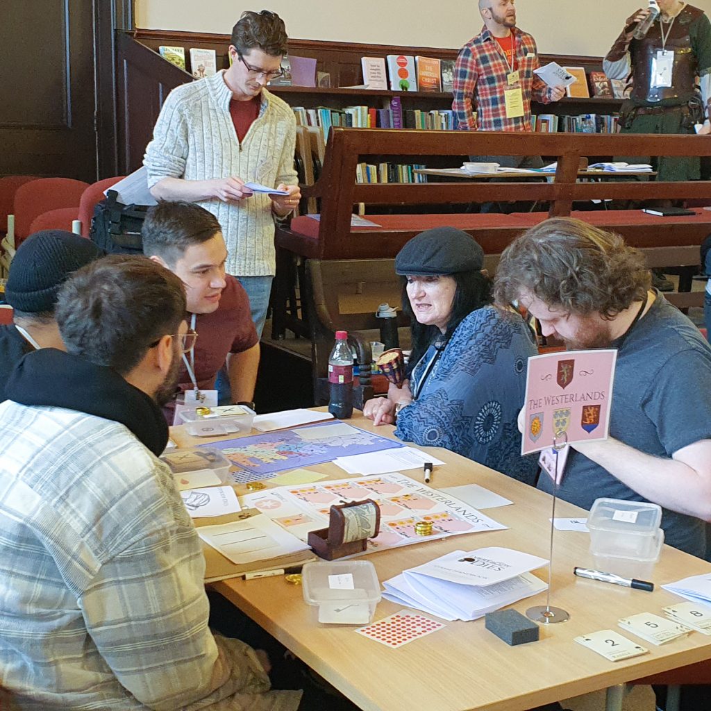 Players deep in discussion around a table with a "The Westerlands" sign. The table is covered in game components.
