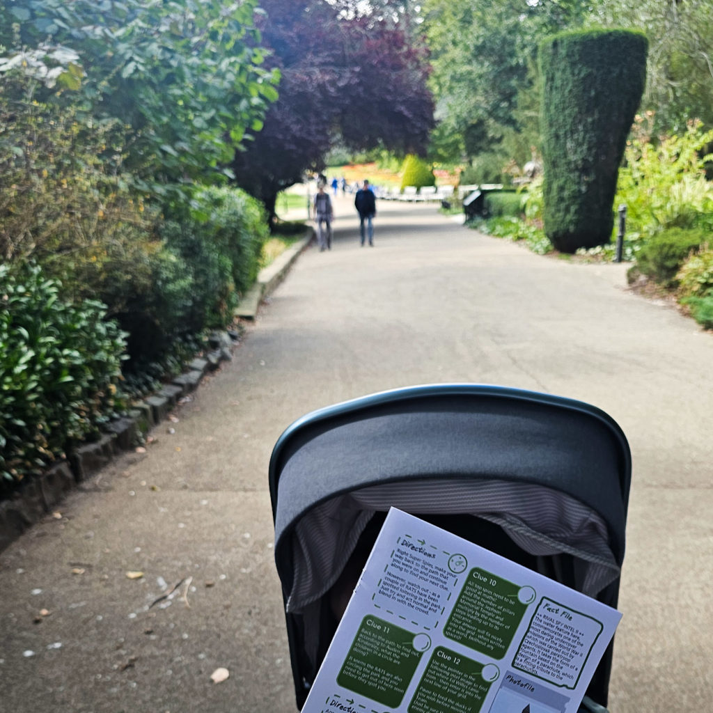 A pram, with the baby inside concealed by the Treasure Trails pack, with a view of a park path