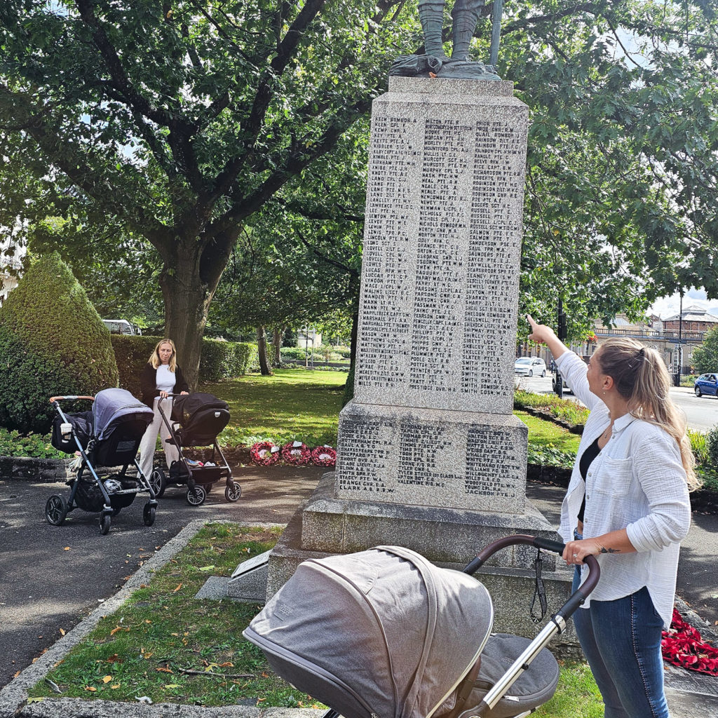 A woman holding a pram and pointing at a memorial, with another woman and two prams visible behind